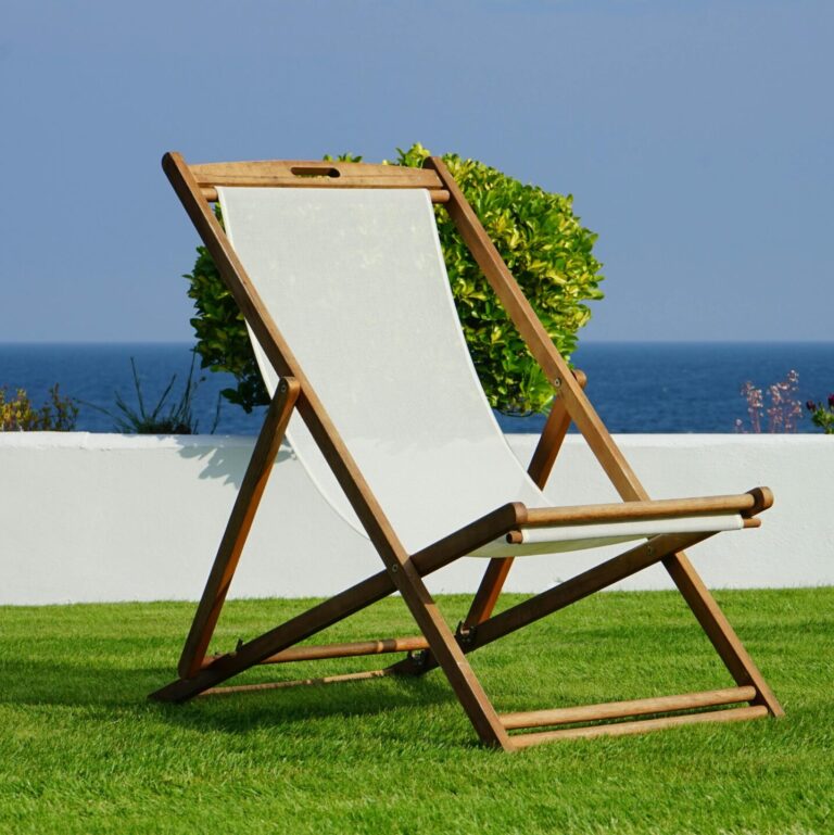A wooden deck chair on lush green grass with a view of the blue ocean, perfect for a summer retreat.